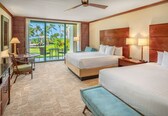 A hotel room with two beds, a desk with a chair, a balcony with a view of palm trees and a pool, and a ceiling fan.