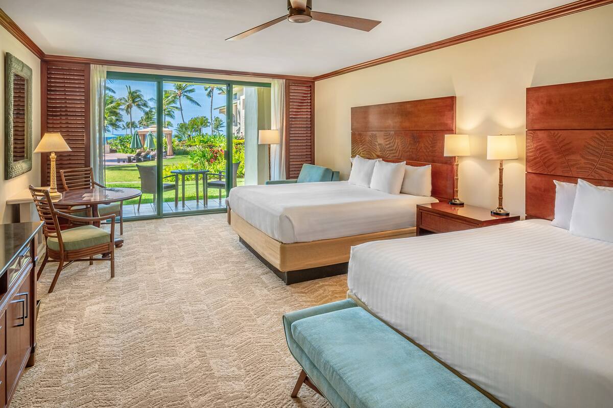 Une chambre d’hôtel avec deux lits, un bureau avec une chaise, un balcon donnant sur des palmiers et une piscine, et un ventilateur de plafond.