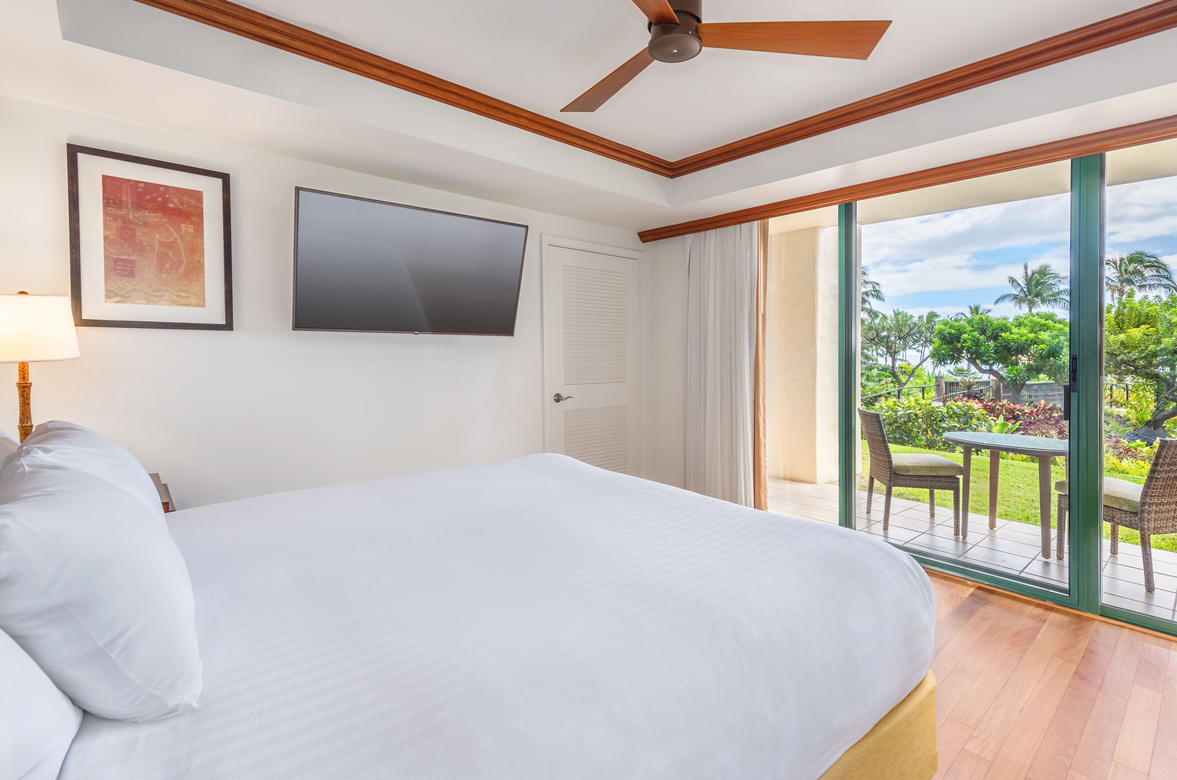 A hotel room with a large bed, a ceiling fan, a flat-screen TV, and a sliding glass door leading to a balcony with outdoor furniture.