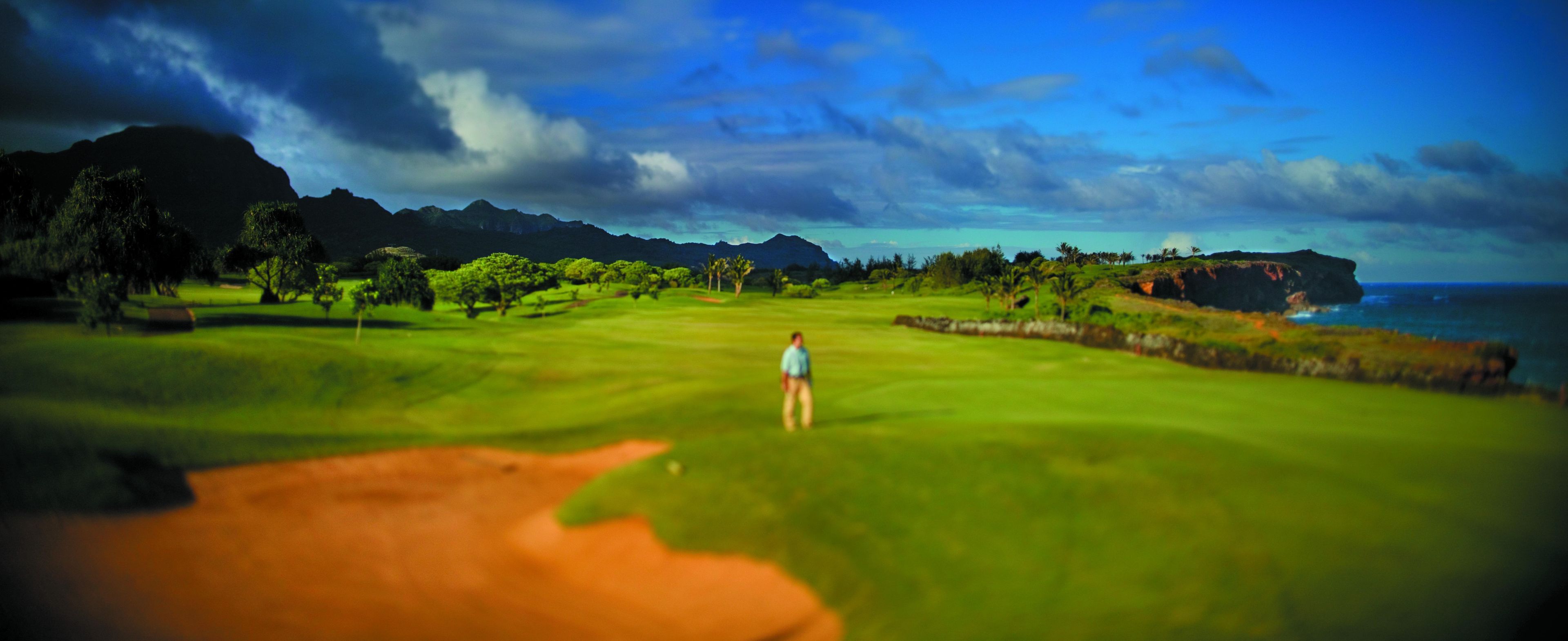 A golf course with a person standing on the green, a sand bunker, and a view of the ocean and mountains in the background.