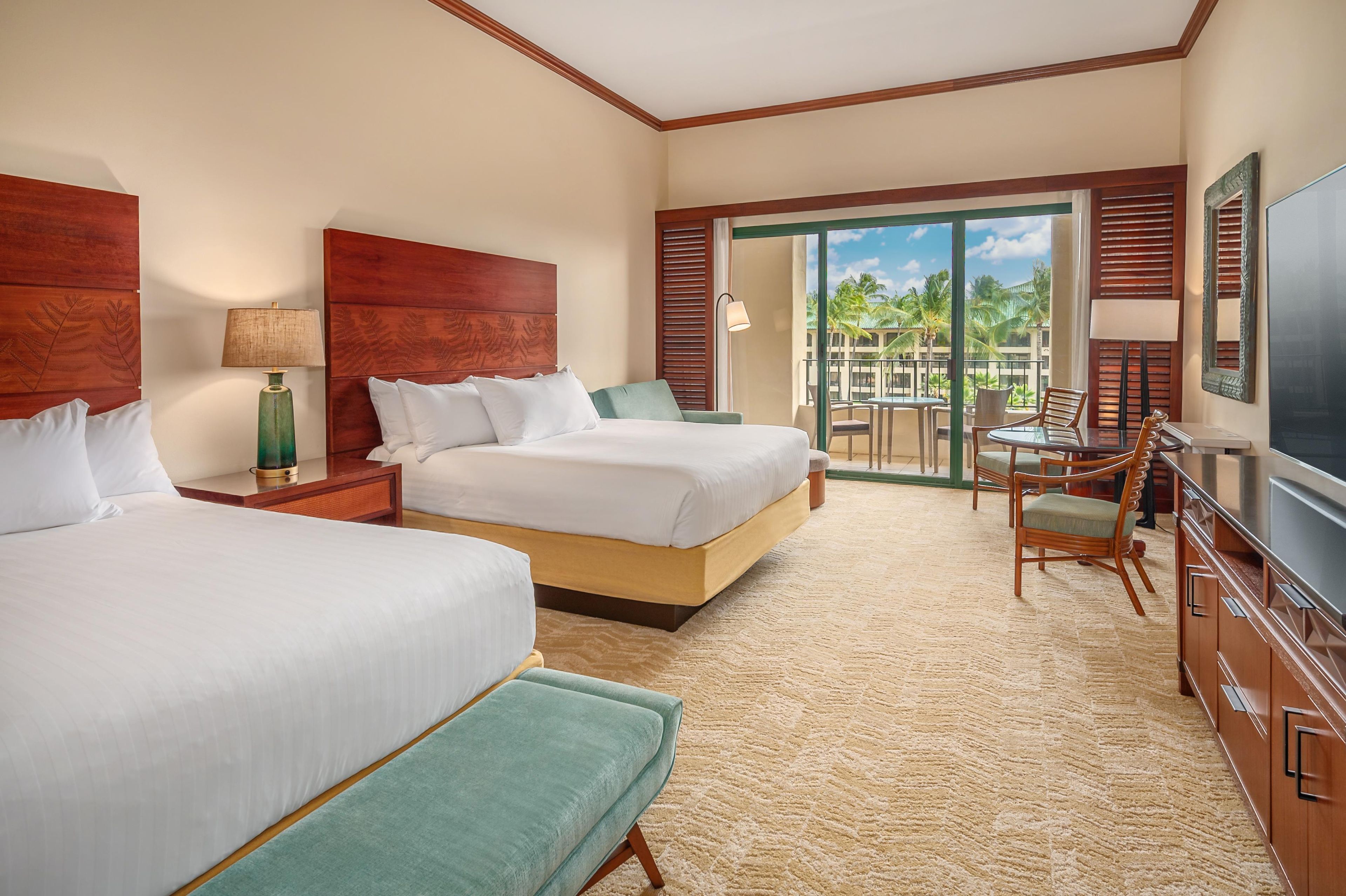 Habitación de hotel con dos camas, televisión, balcón con vistas a palmeras y zona de comedor con sillas y mesa.