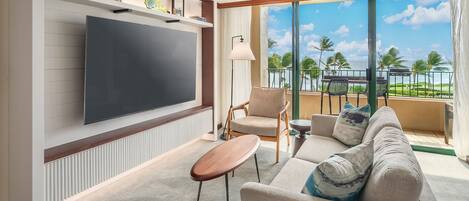 Una sala de estar moderna con un televisor de pantalla plana grande, un sofá, una mesa de centro y un balcón con vistas a palmeras y al océano.