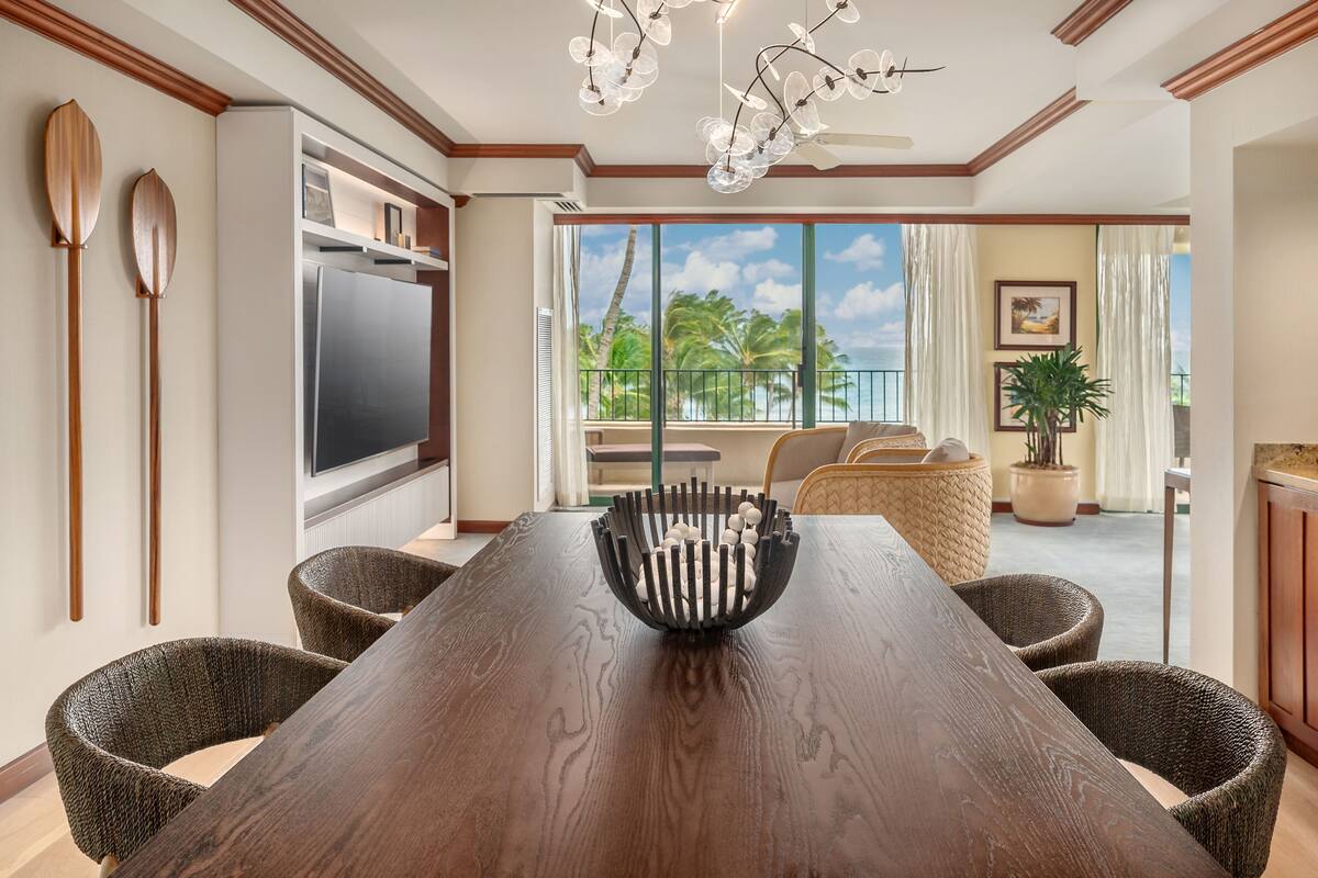 Un coin repas avec une table en bois, des chaises en osier, un lustre moderne et une vue sur l’extérieur.