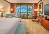 A hotel room with a bed, a desk, a TV, a balcony with a view of palm trees and the ocean.