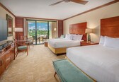 A hotel room with two beds, a desk with a chair, a TV, and a balcony with a view.