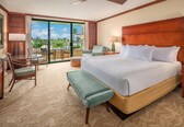 A hotel room with a large bed, a seating area with a table and chairs, a balcony with a view, and a wooden headboard.
