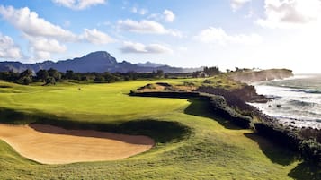 Un campo de golf con búnkeres, una bandera y vistas al océano y las montañas.