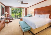 A hotel room with a large bed, a desk with a chair, a bench, a ceiling fan, and a view of the outdoors.