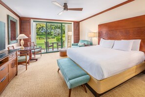 A hotel room with a large bed, a desk with a chair, a bench, a ceiling fan, and a view of the outdoors.