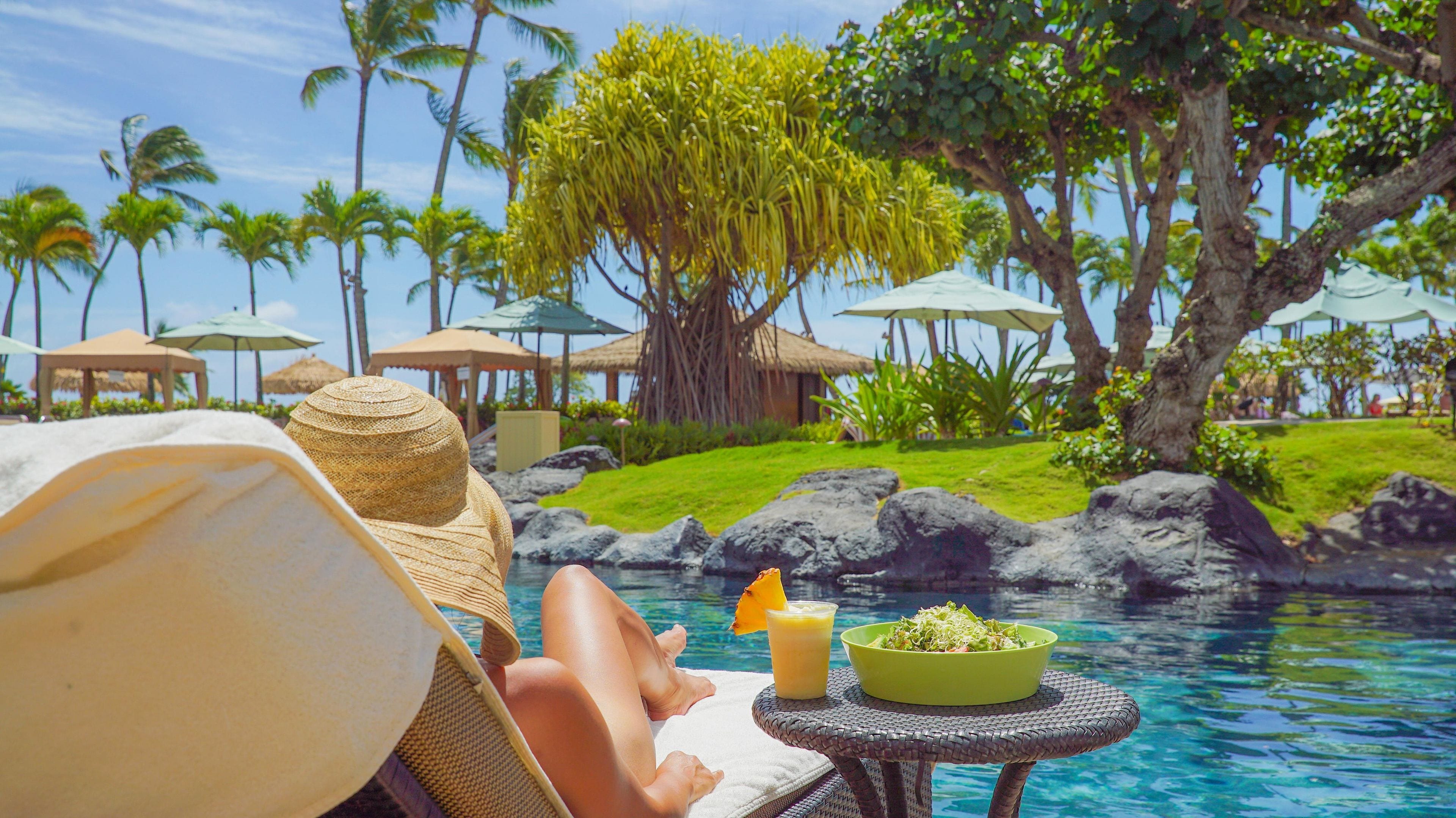 A poolside view with lounge chairs, a dining table with a bowl of salad and a drink, and tropical landscaping.