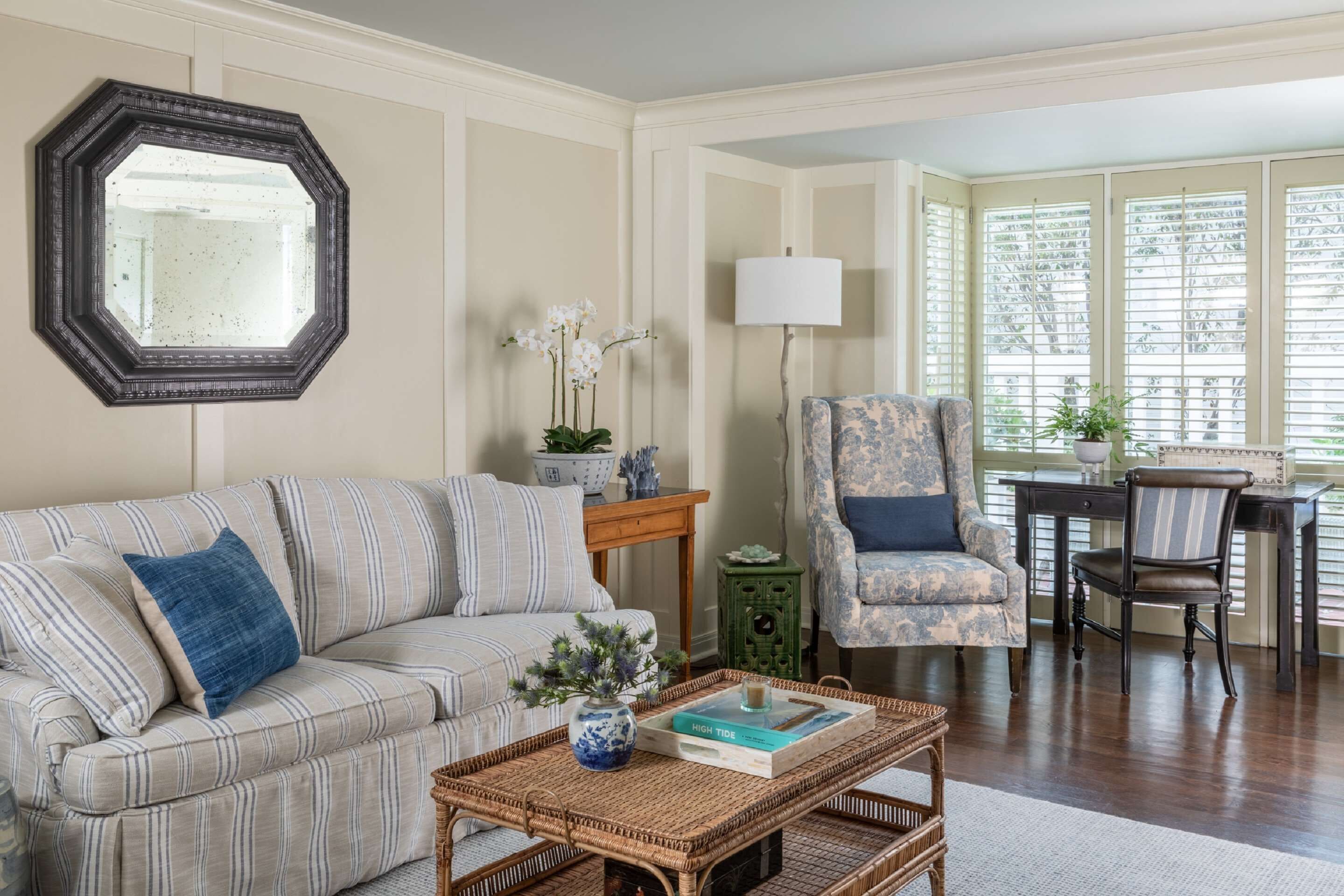 A living room with a striped sofa, a coffee table with a book and vase, a chair, a lamp, and a large window with blinds.