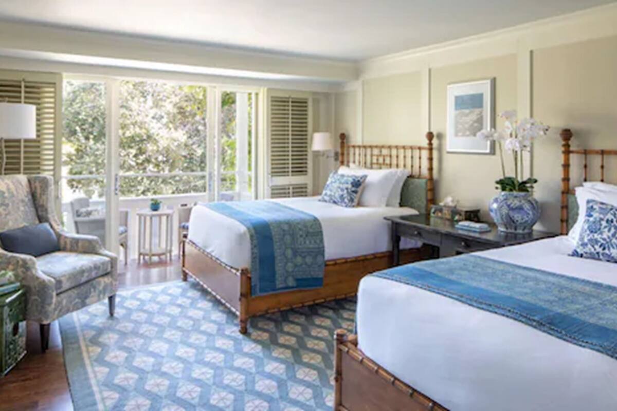 A hotel room with two beds, a chair, a small table, and a window with shutters.