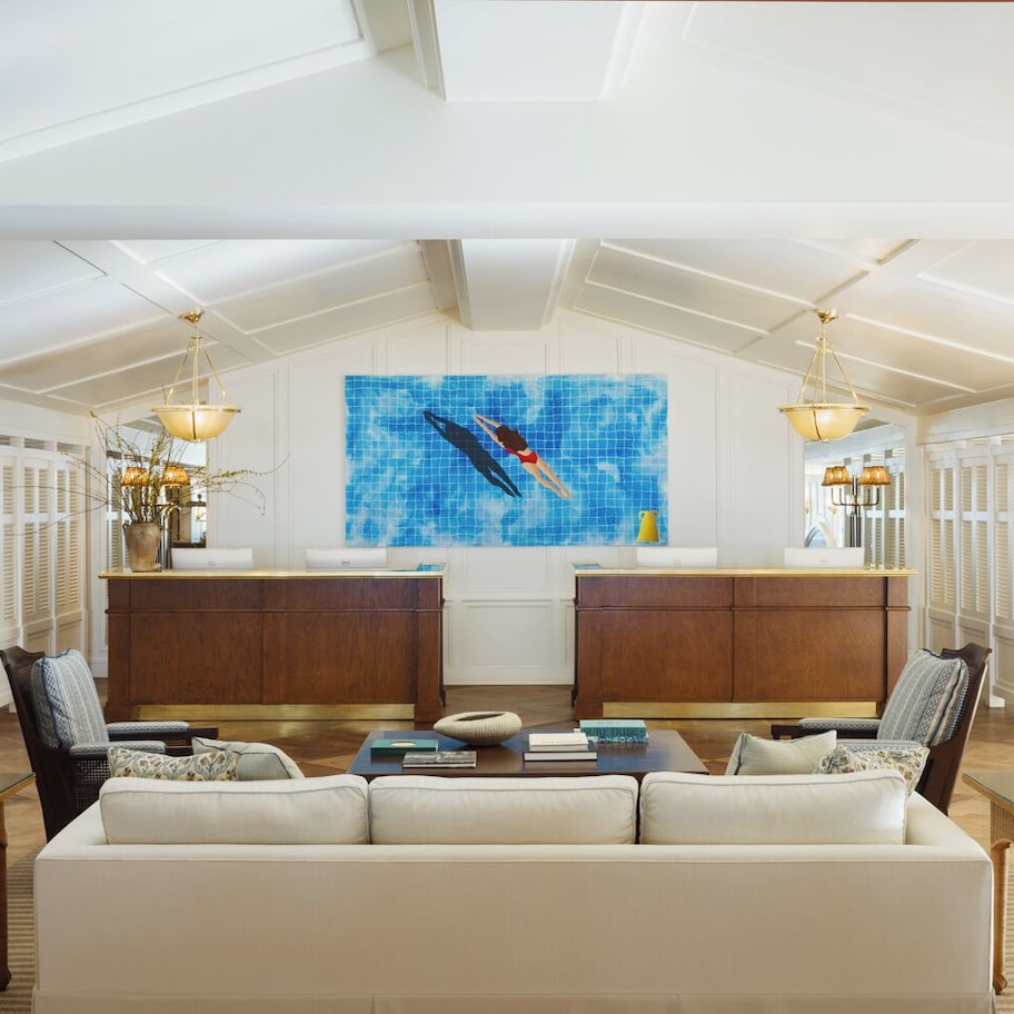 A spacious living room with a high ceiling, a central seating area, a kitchen with a wooden cabinet, and a large blue mosaic artwork on the wall.