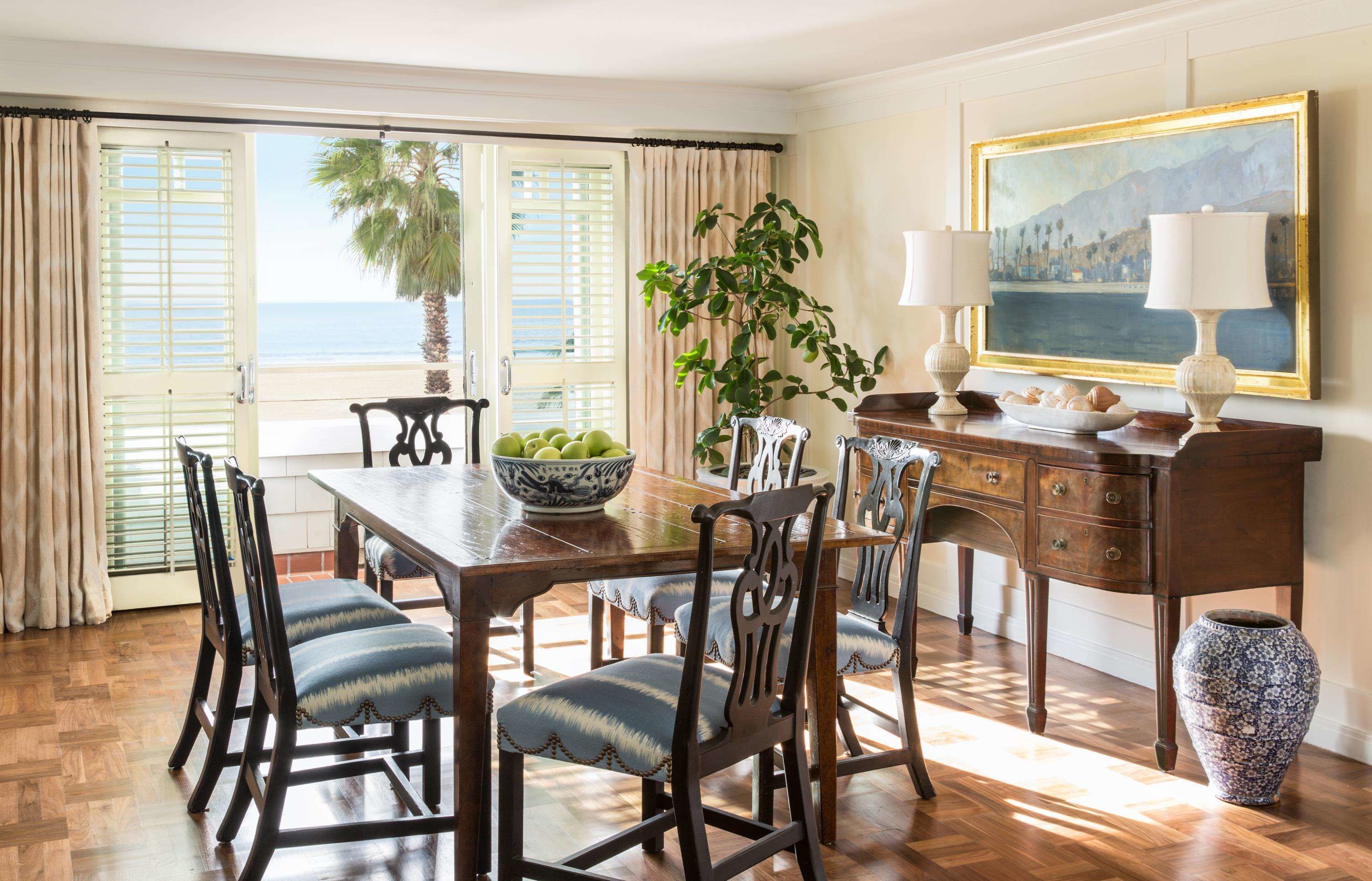 A dining area with a wooden table, chairs, a bowl of fruit, a potted plant, and a painting on the wall.