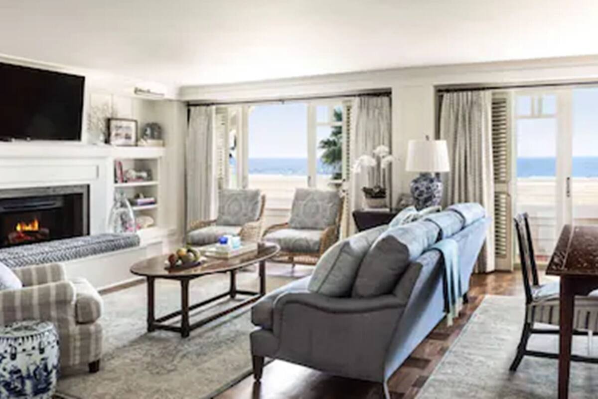A spacious living room with a fireplace, comfortable seating, a coffee table, and large windows offering a view of the beach.