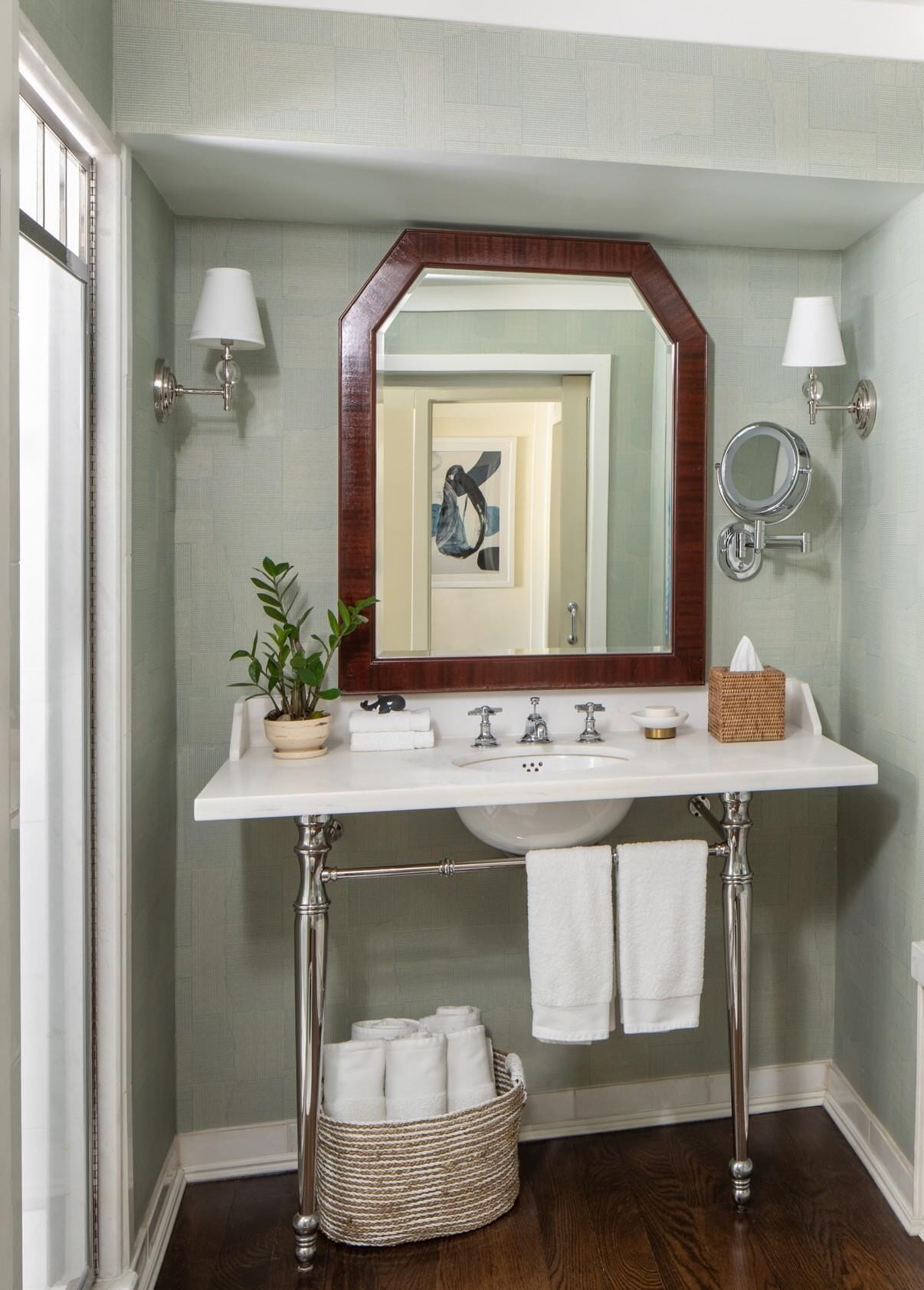A bathroom with a white vanity, a mirror, two wall-mounted lights, a potted plant, a towel rack, and a wicker basket.