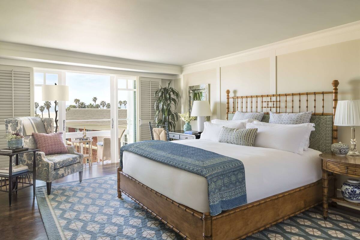 A spacious bedroom with a large bed, a chair, a desk, and a view of palm trees through the window.