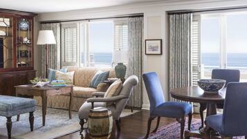 A well-furnished living room with a sofa, armchairs, a coffee table, and a dining table with chairs. There is a large window with a view of the ocean.