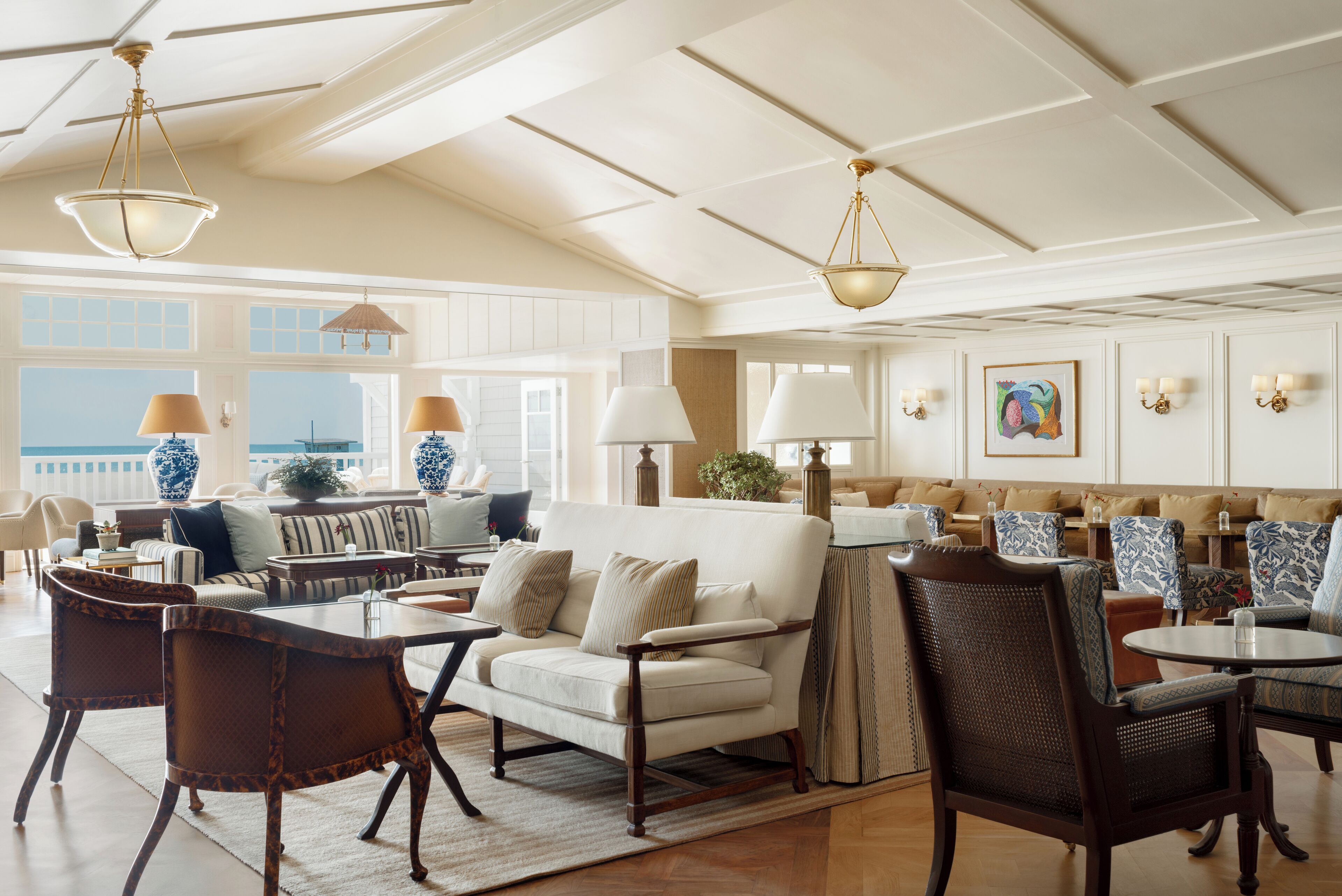 A spacious living area with a high ceiling, large windows, and a view of the ocean. There are multiple seating areas with tables and chairs.