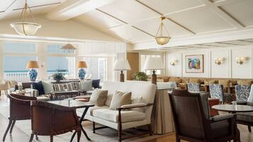 A spacious living area with a high ceiling, large windows, and a view of the ocean. There are multiple seating areas with tables and chairs.