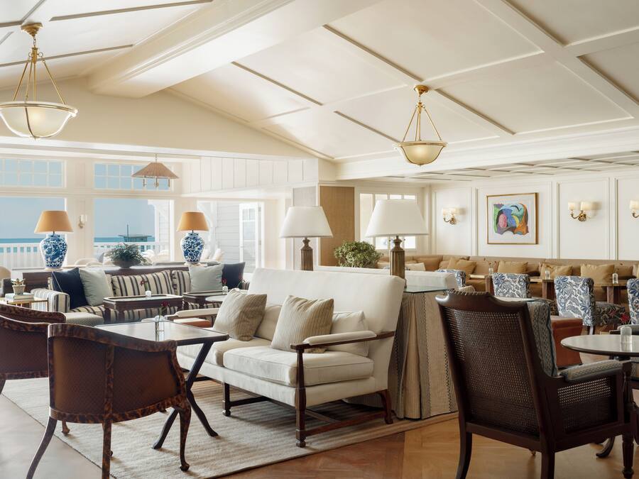 A spacious living area with a high ceiling, large windows, and a view of the ocean. There are multiple seating areas with tables and chairs.