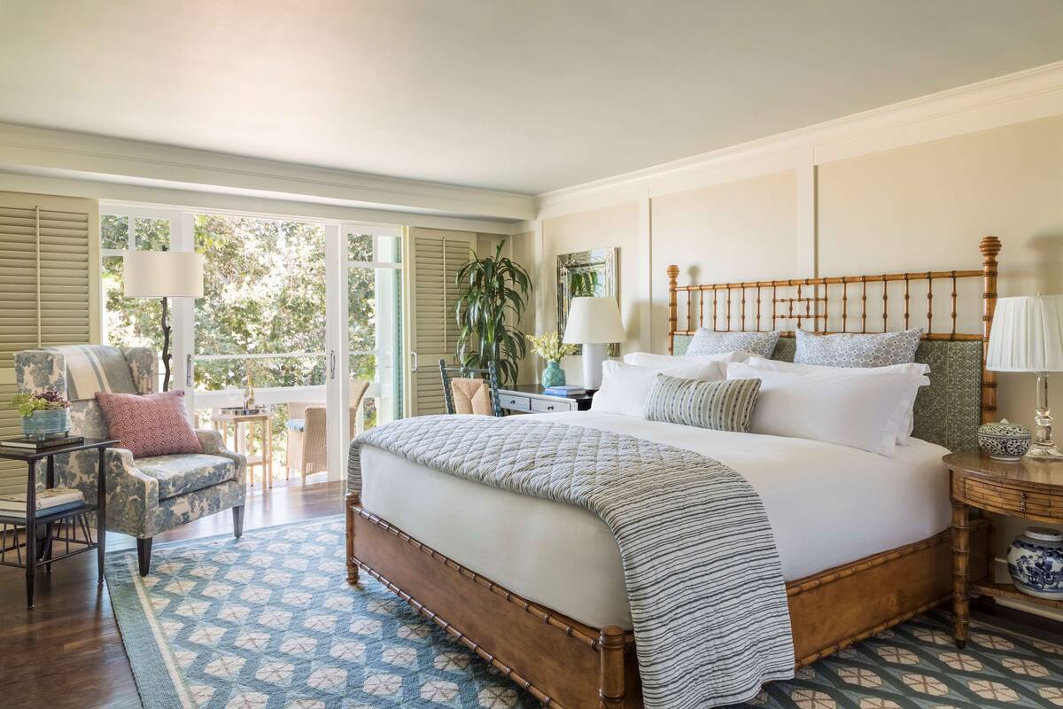 A bedroom with a large bed, a chair, a small table, and a window with a view of trees.