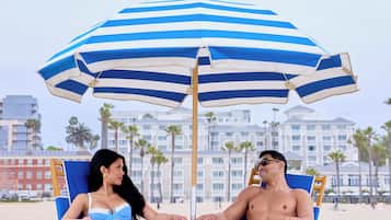 Two people sitting under a striped beach umbrella on a sandy beach with palm trees and hotel buildings in the background.