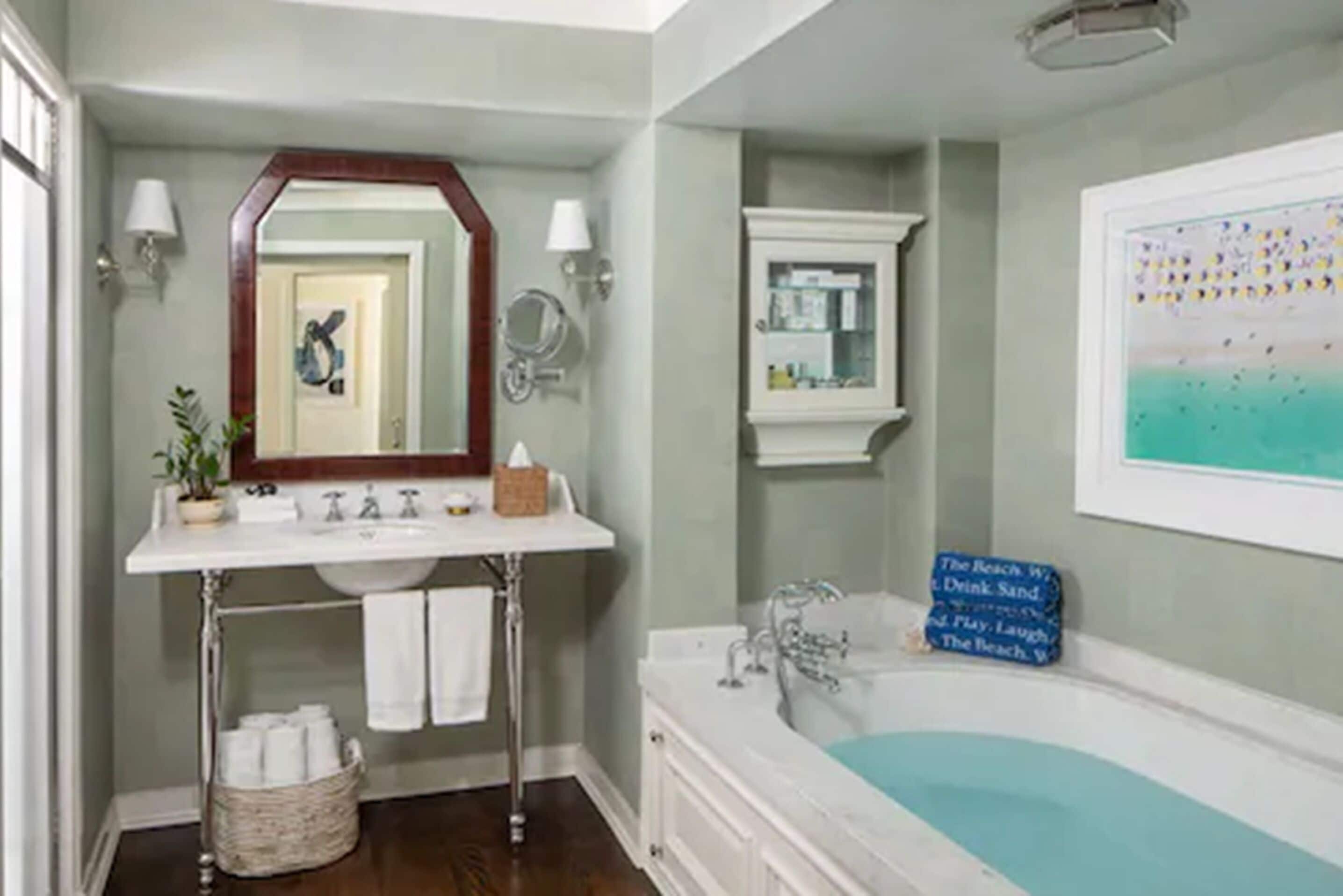 A bathroom with a white sink, mirror, and bathtub. There is a towel rack, a basket of toiletries, and a framed picture on the wall.