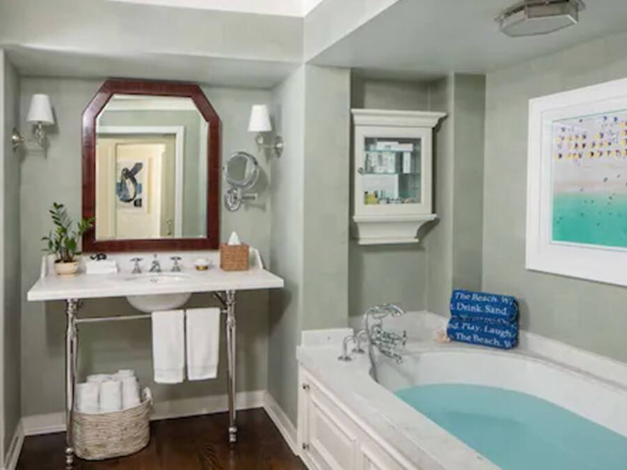 A bathroom with a white sink, mirror, and bathtub. There is a towel rack, a basket of toiletries, and a framed picture on the wall.