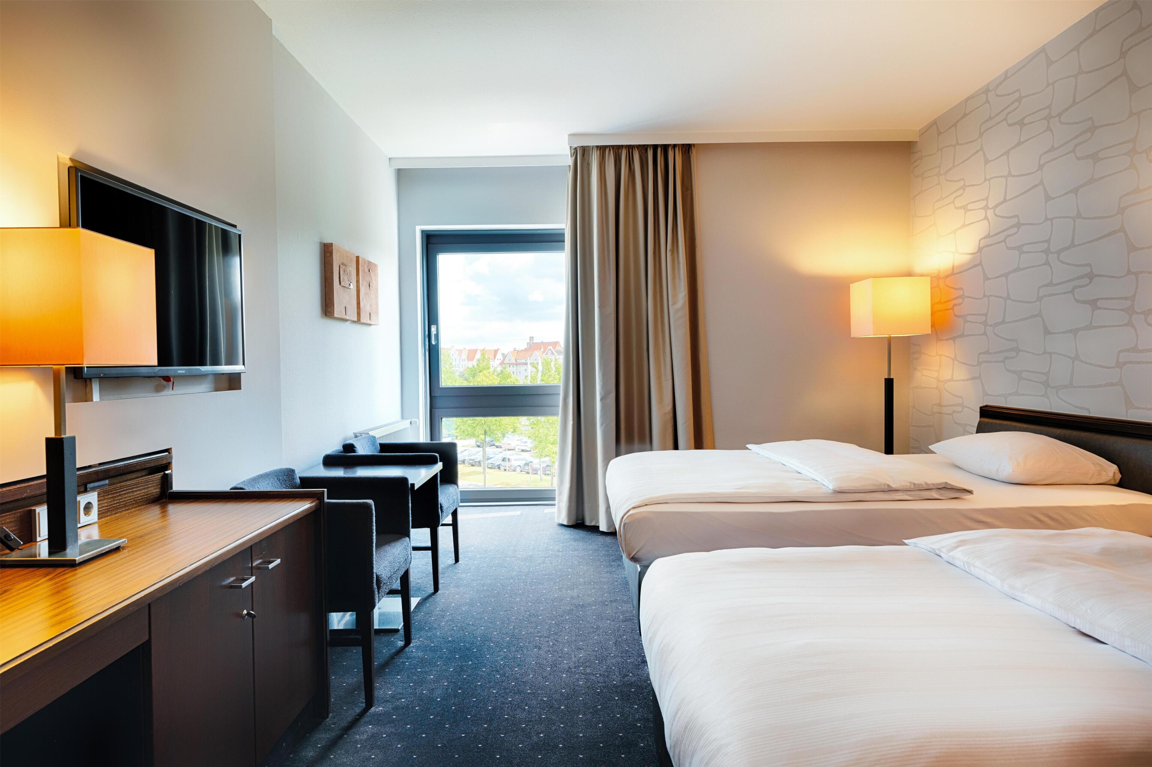 A hotel room with a desk, two beds, a TV, and a window with curtains.