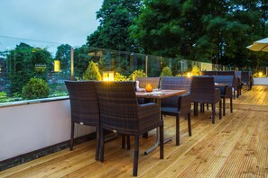 An outdoor dining area with wicker chairs and tables, a glass railing, and a view of greenery and buildings. - Park Inn by Radisson Lübeck (Lübeck)