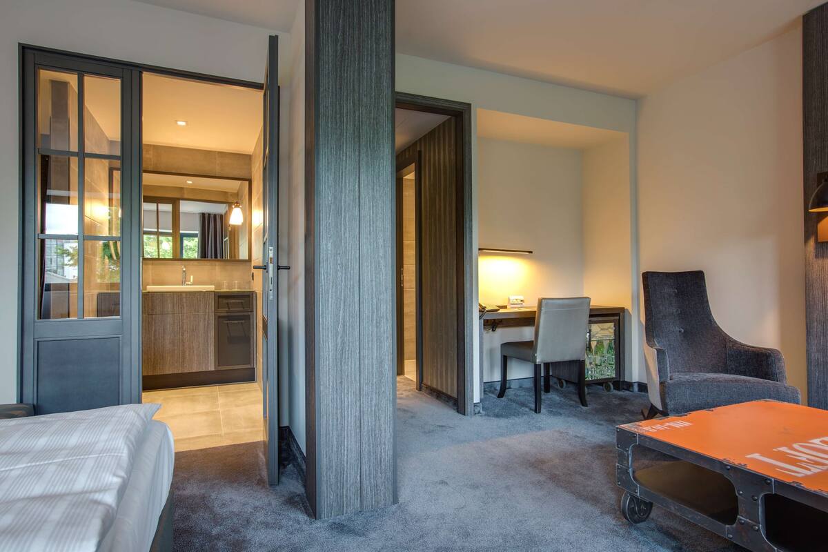 Habitación de hotel con cama, escritorio, silla y mesa de comedor. Se ve un área de cocina a través de una puerta abierta.