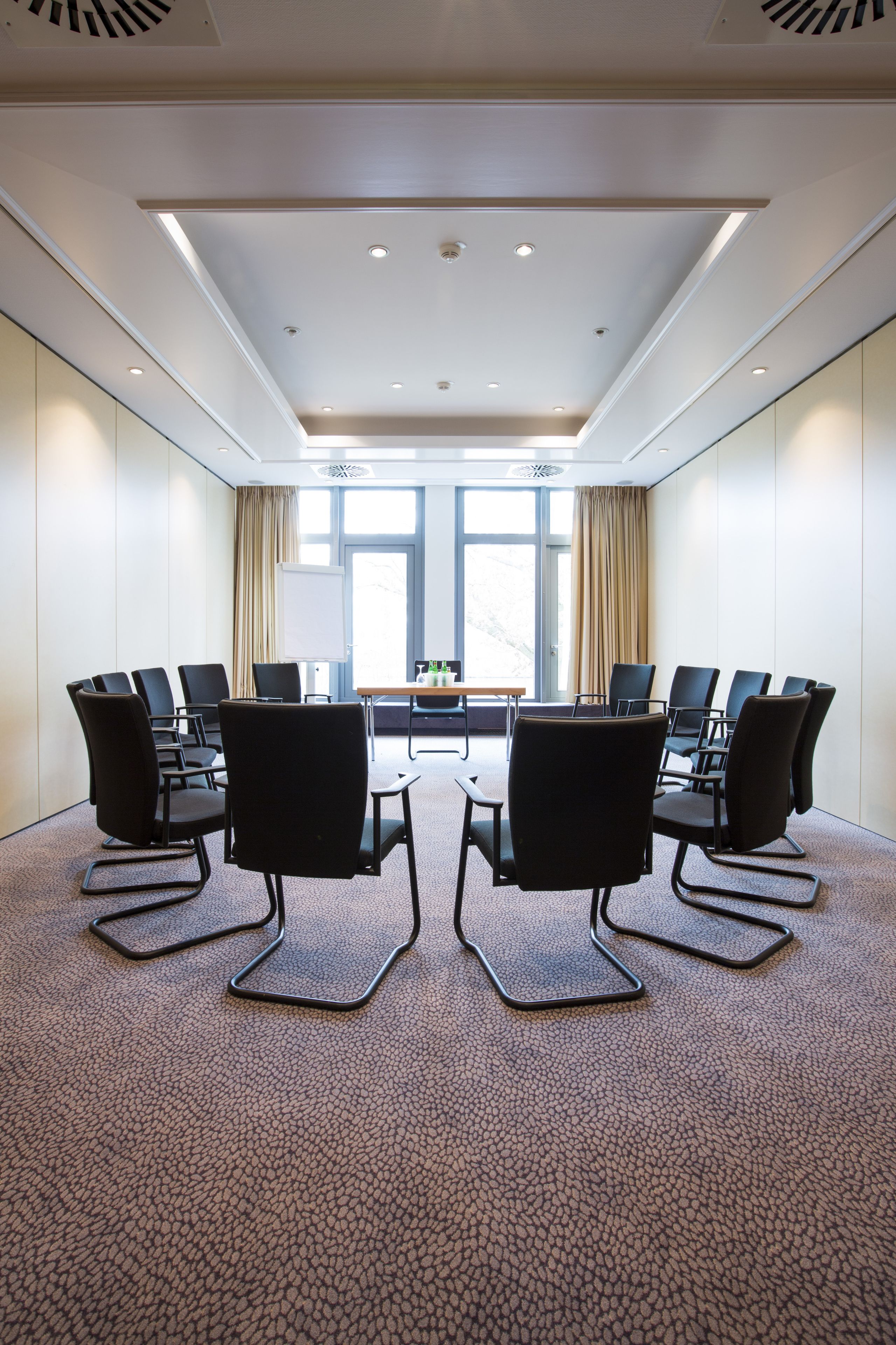 A conference room with a rectangular table surrounded by eight chairs, large windows with curtains, and a carpeted floor.