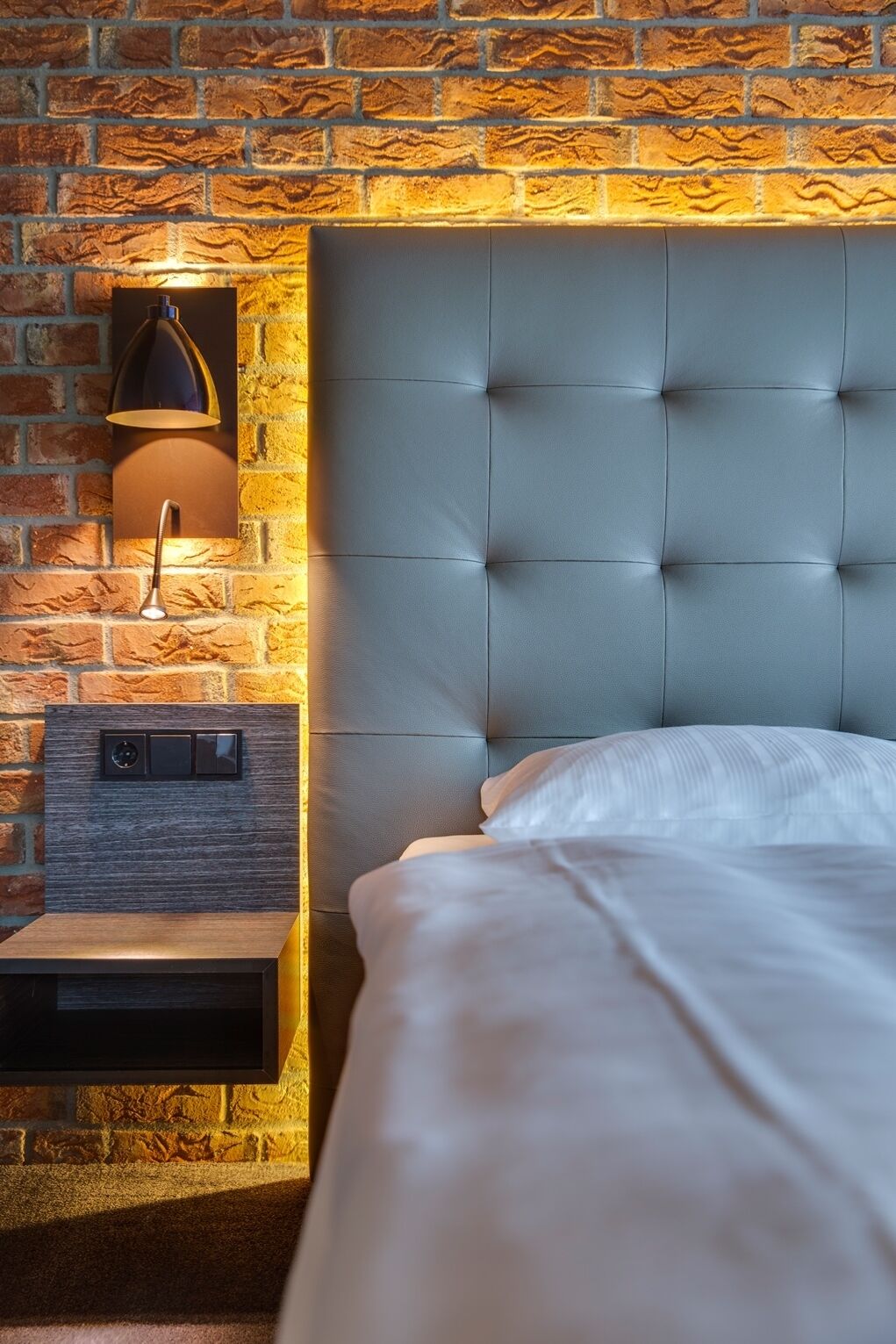 Une chambre avec un mur en briques, une tête de lit capitonnée, une table de chevet avec une lampe et un lit avec une literie blanche.