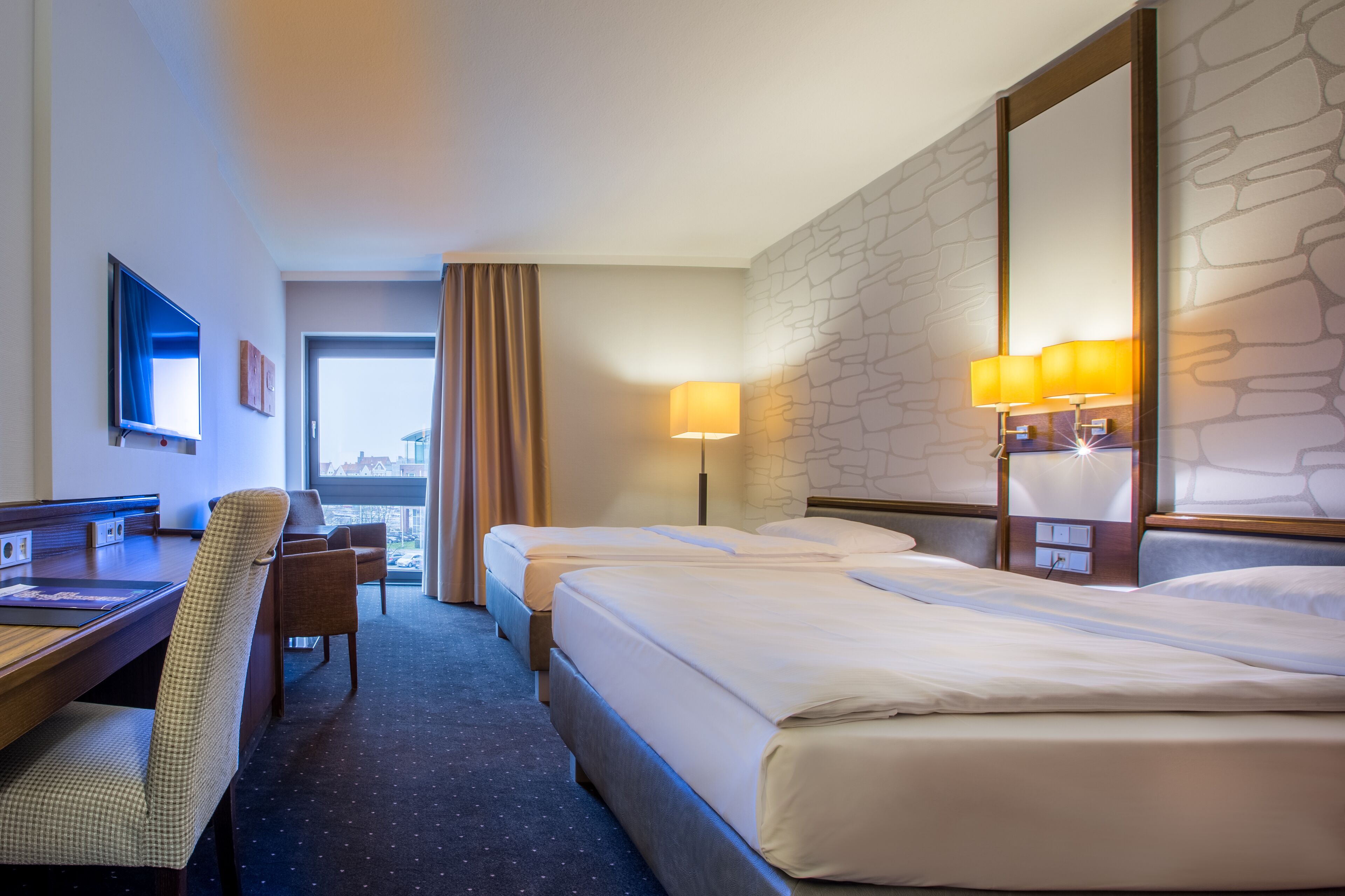 Une chambre d’hôtel avec deux lits, un bureau avec une chaise, une télévision et une grande fenêtre avec des rideaux.