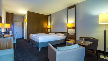 A hotel room with a large bed, a desk with a lamp, a chair, and a small table with a glass.
