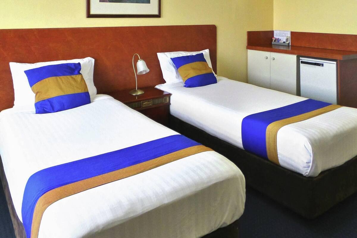 Dos camas individuales con sábanas blancas, detalles en azul y dorado, una mesita de noche con lámpara y un refrigerador pequeño.