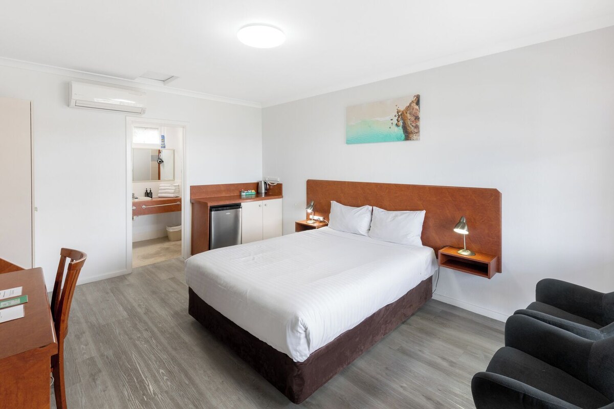 A hotel room with a large bed, a desk with a chair, a small kitchenette, and a bathroom visible through an open door.