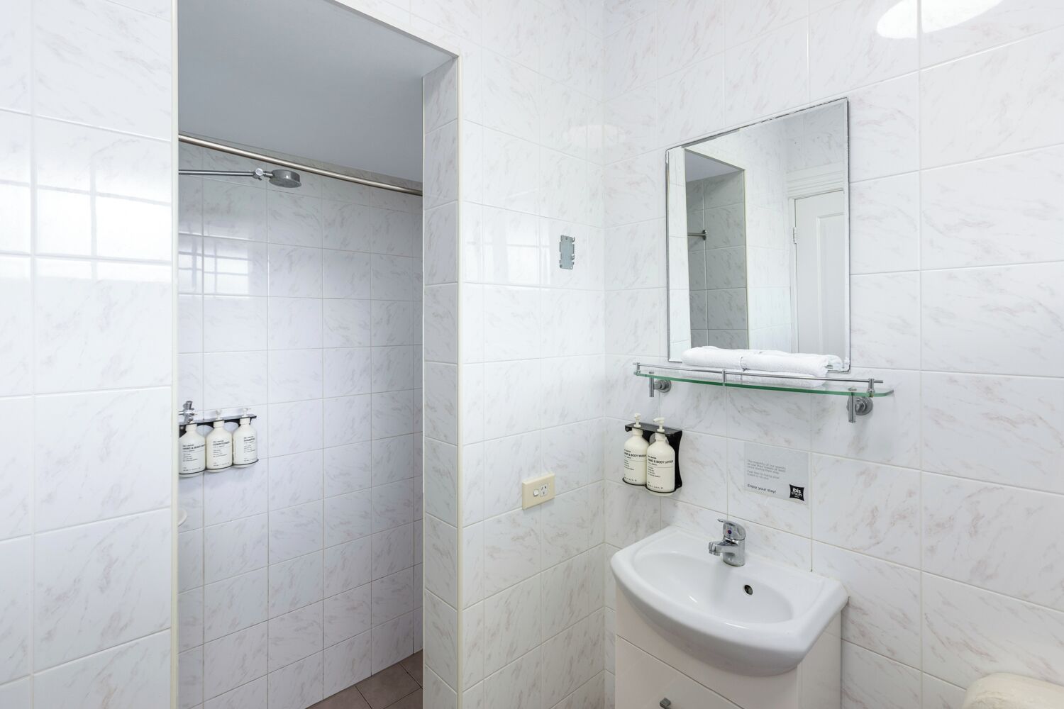 Un baño moderno con baldosas de mármol blanco, cabina de ducha de vidrio, lavamanos blanco y espejo sobre el lavamanos.