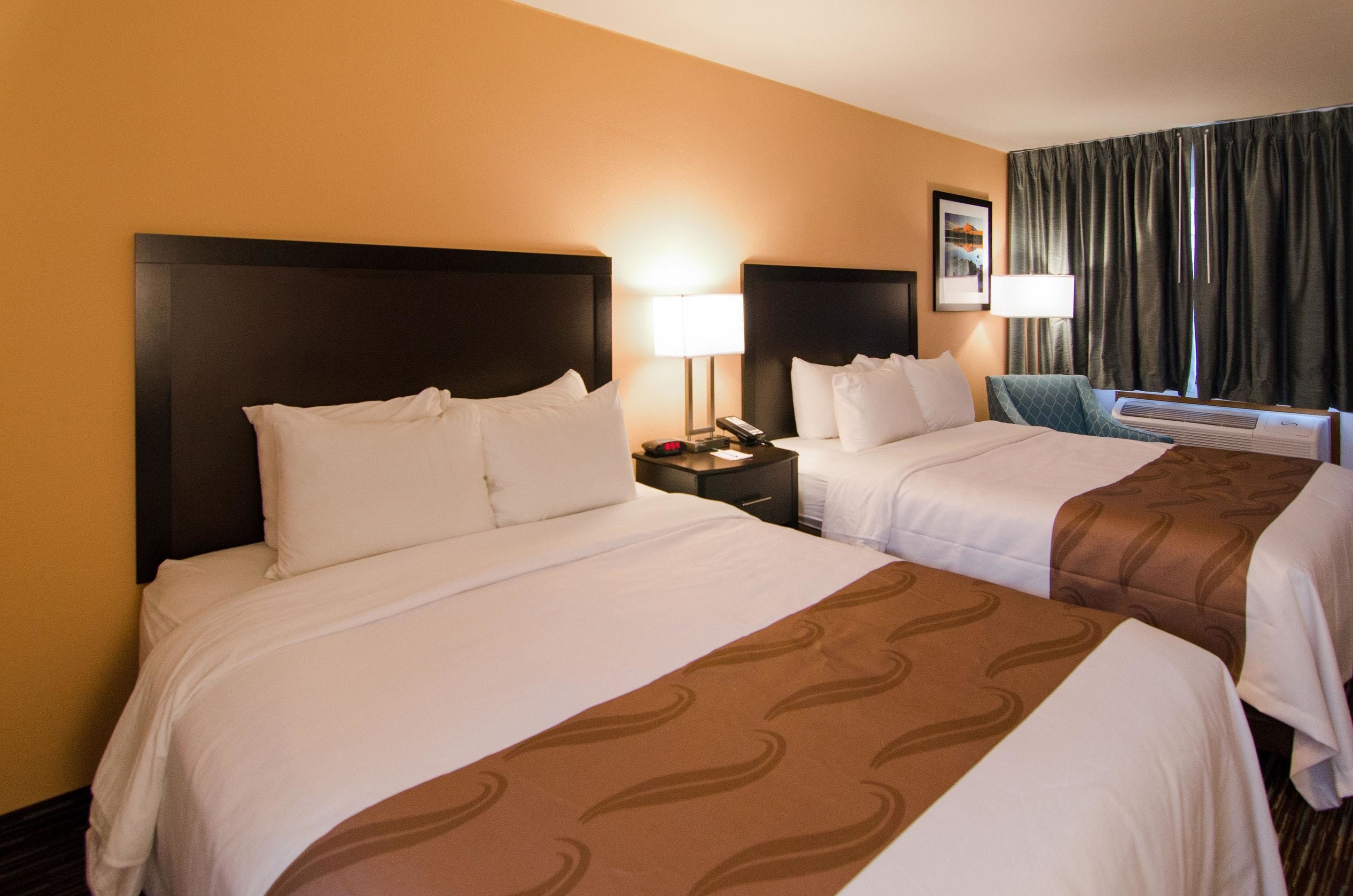 A hotel room with two beds, a nightstand with a lamp, and a window with curtains.