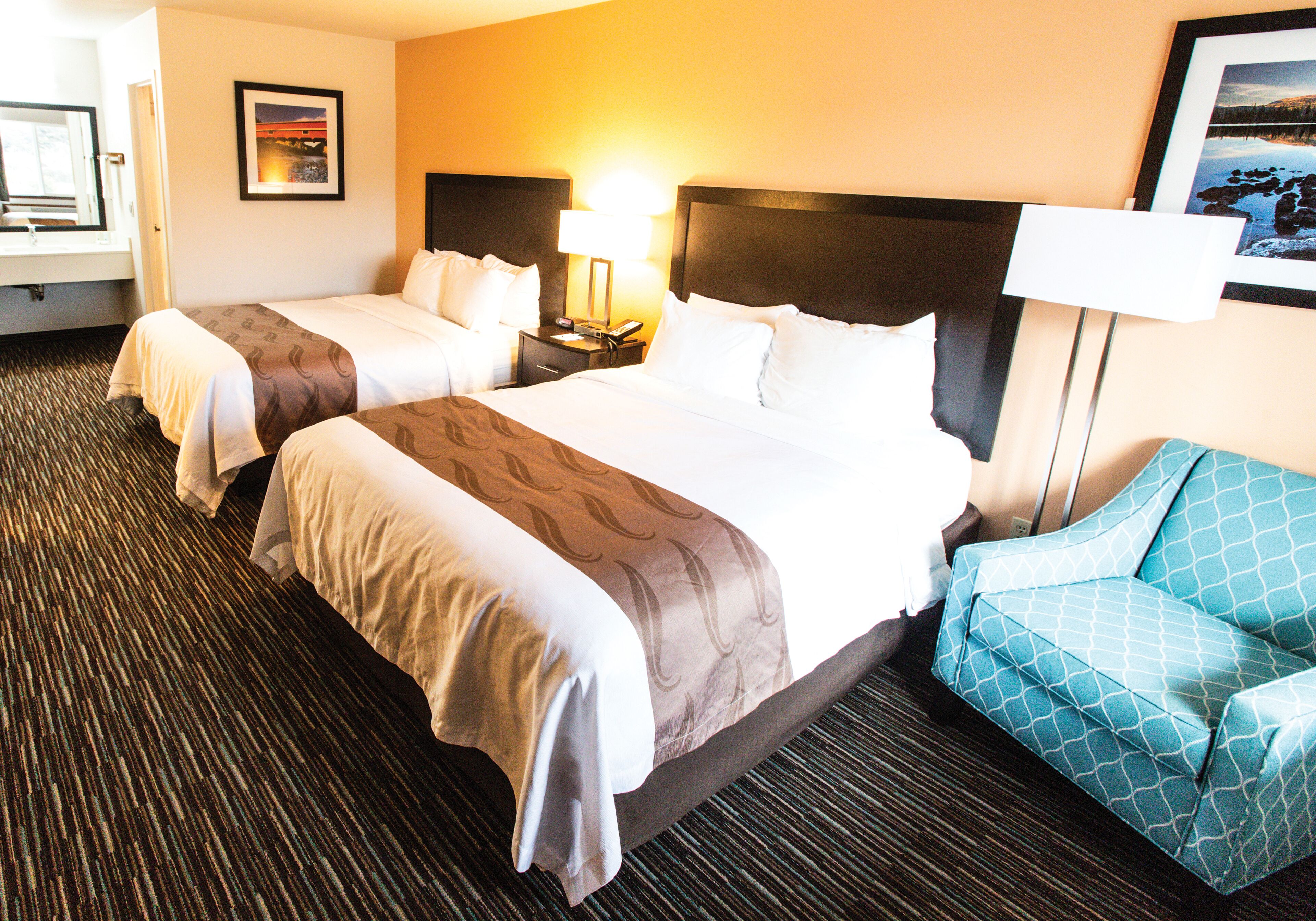 A hotel room with two beds, a nightstand with a lamp, a chair, and a framed picture on the wall.