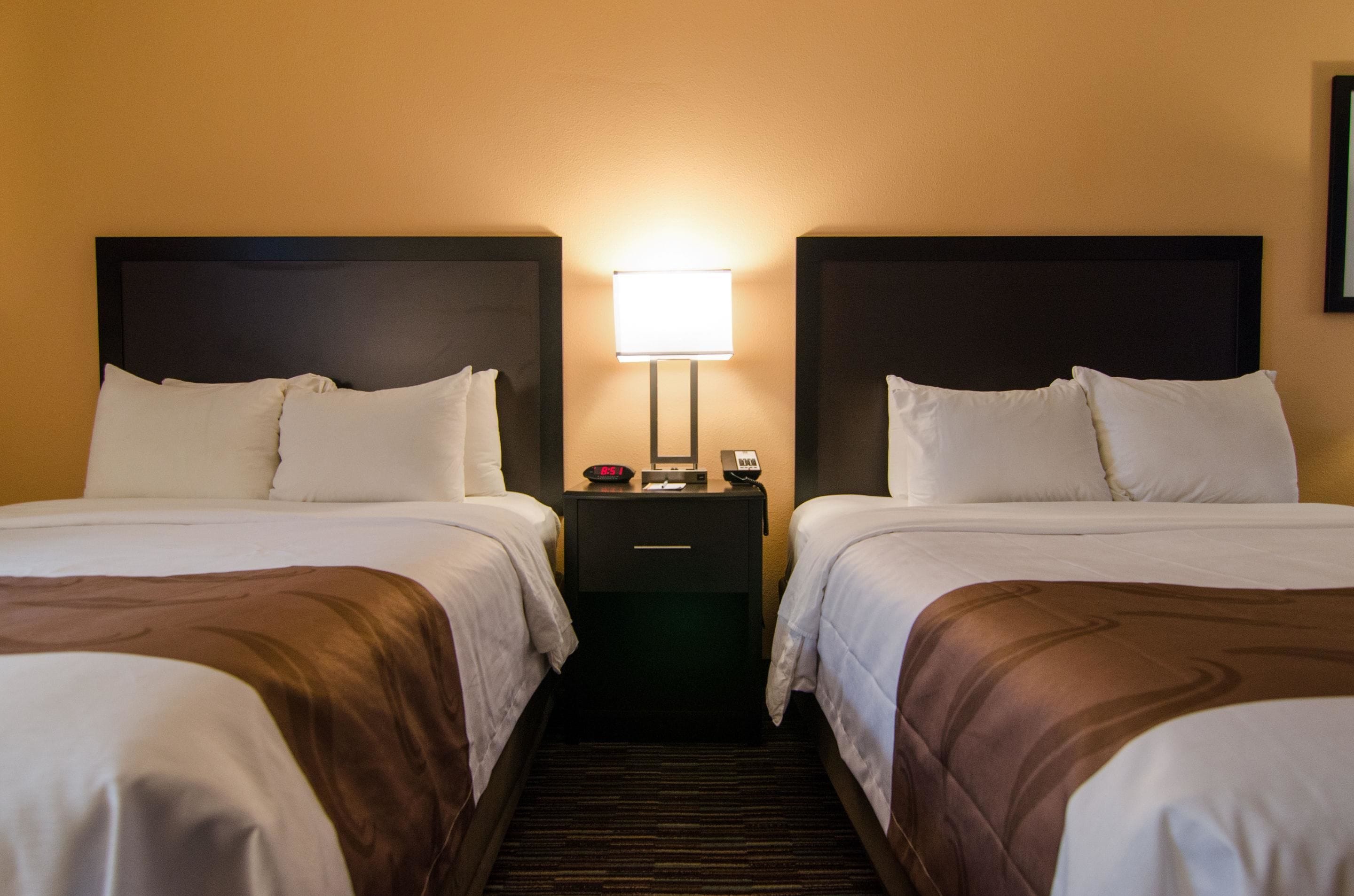 A hotel room with two beds, a nightstand with a lamp, and a wall-mounted phone.
