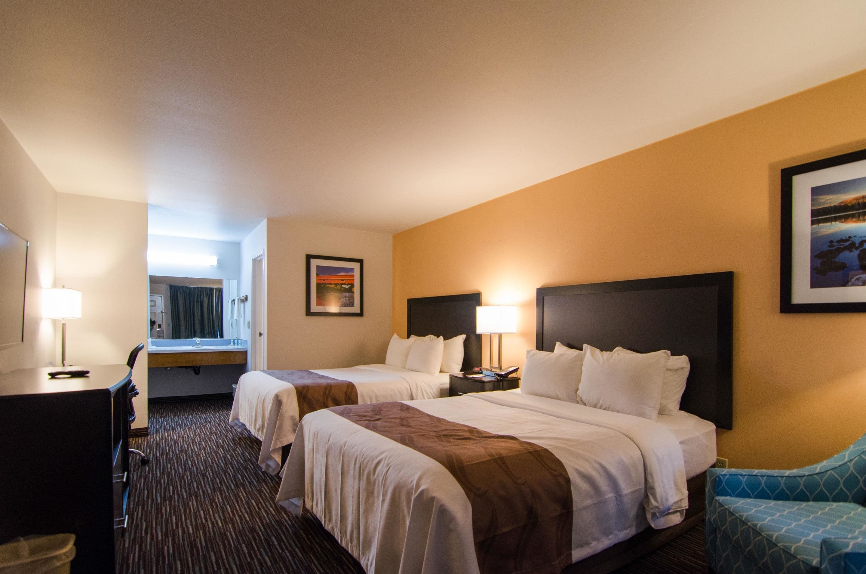 A hotel room with two beds, a desk, a chair, a bathroom, and a view of the outdoors.