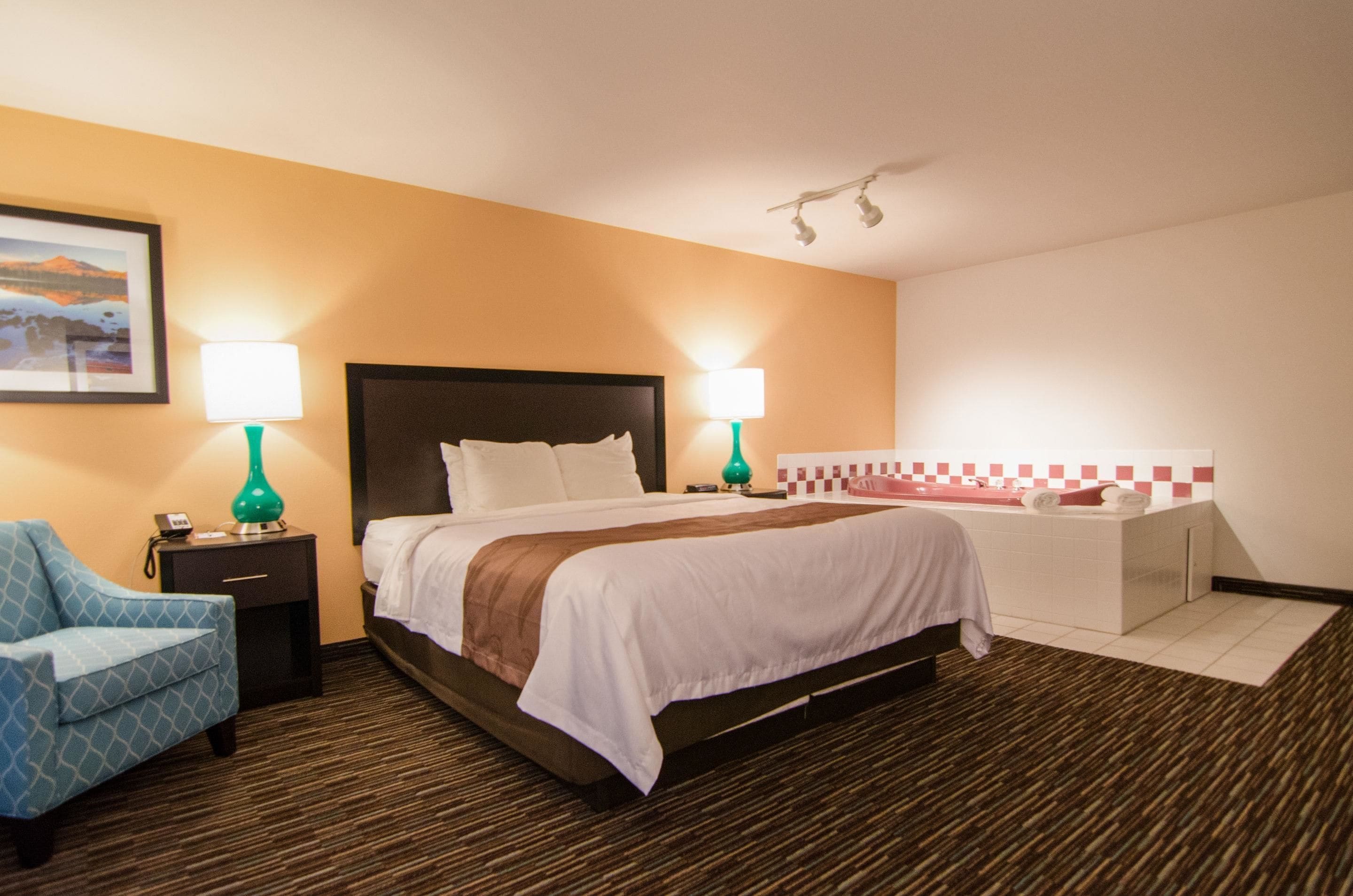 A hotel room with a bed, two bedside tables with lamps, a chair, a bathroom with a bathtub, and a wall-mounted light fixture.