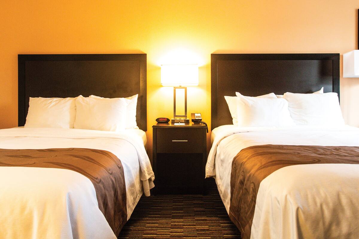A hotel room with two beds, a nightstand with a lamp, and a carpeted floor.