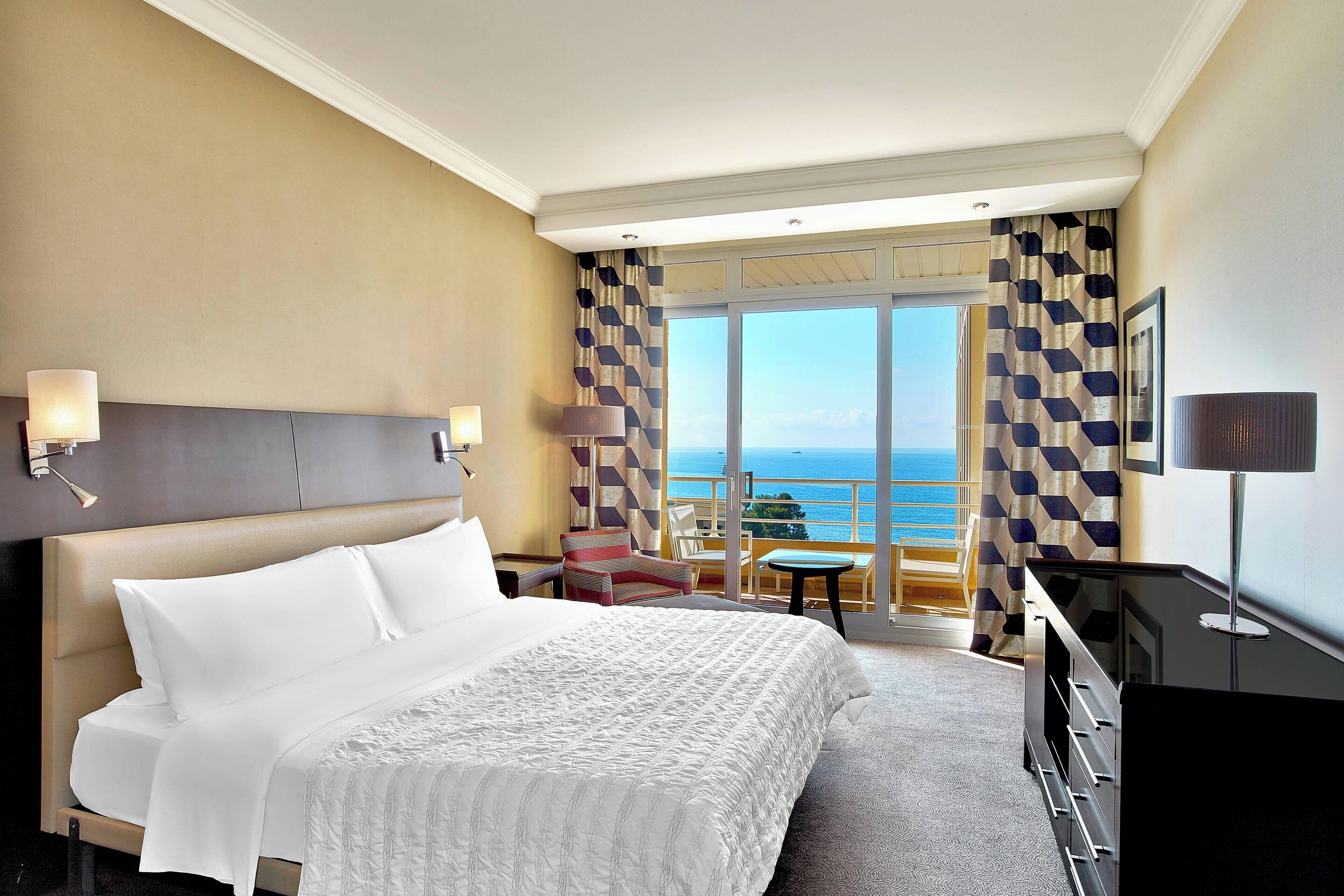 A hotel room with a large bed, a desk, a chair, a TV, and a balcony with a view of the sea.