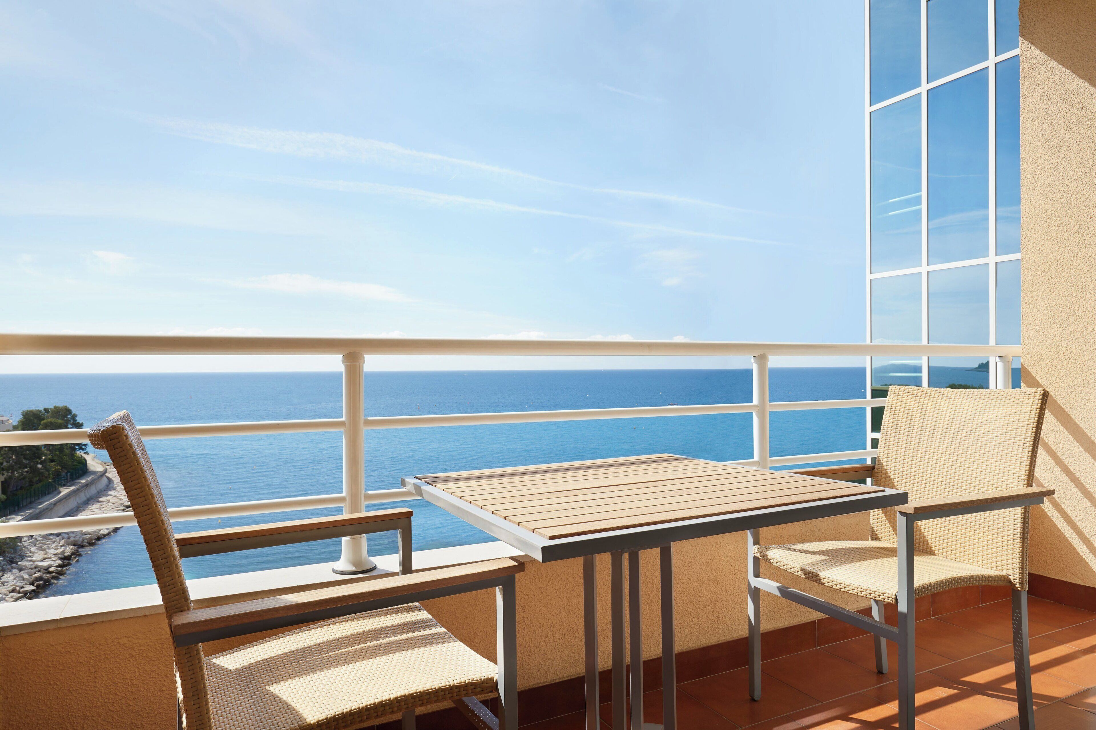 Een balkon met een tafel en twee rieten stoelen, met uitzicht op de kust.