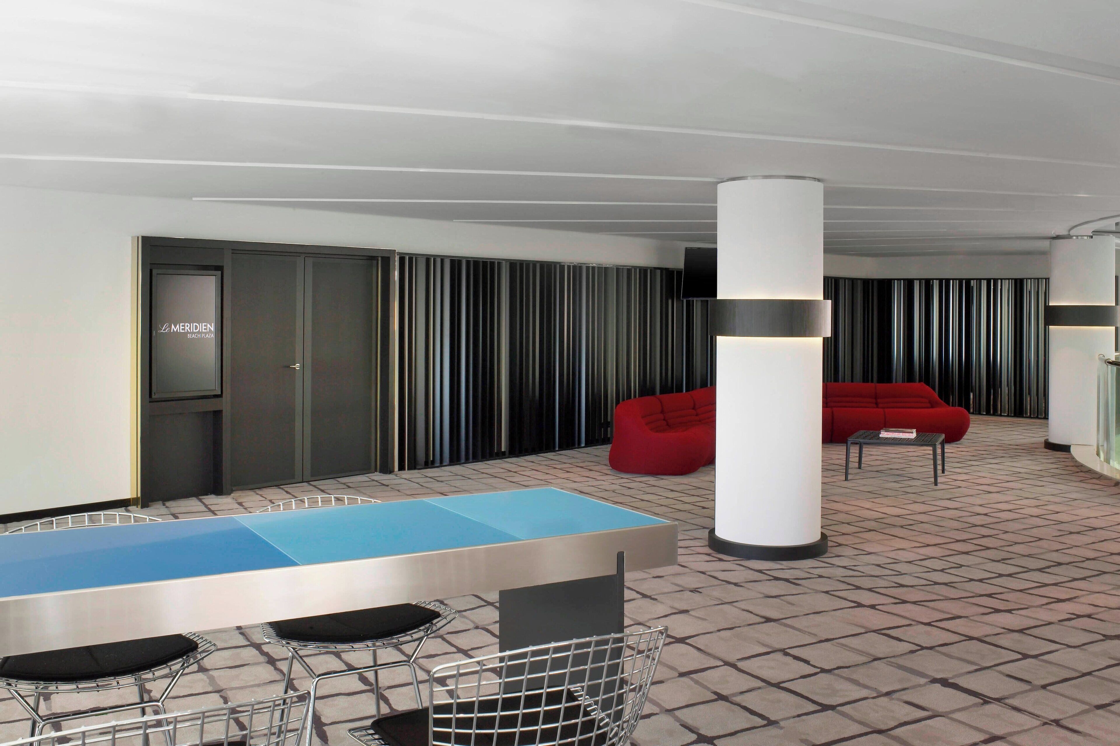 A modern lounge area with a blue and white table, metal chairs, a red sofa, and a patterned floor.