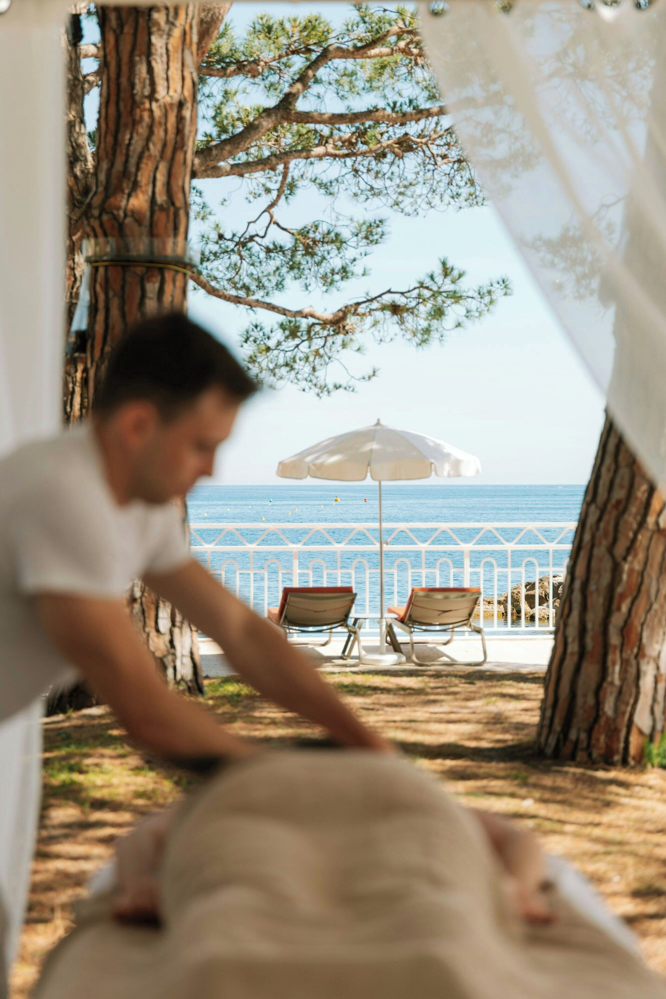 Une personne penchée au-dessus d’un fauteuil inclinable, avec une vue sur la mer et un parasol blanc en arrière-plan.