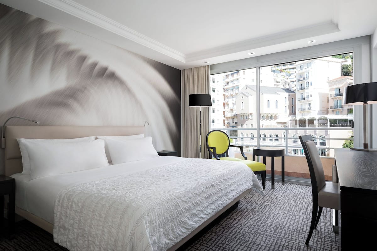 A hotel room with a large bed, a desk, a chair, and a view of the city through large windows.